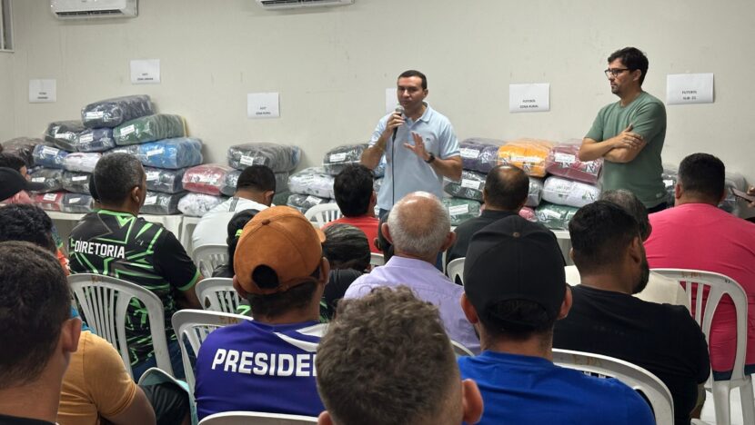 Emídio Júnior fala ao microfone diante de representantes esportivos, com conjuntos de uniformes embalados ao fundo