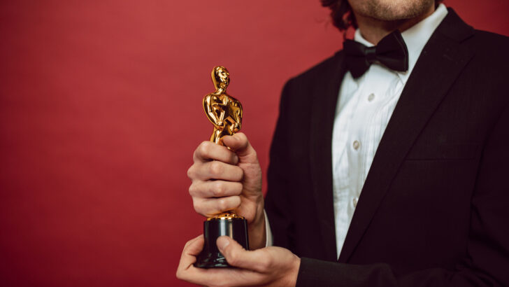 Homem de smoking segura uma estatueta dourada de premiação diante de fundo vermelho, em imagem ilustrativa