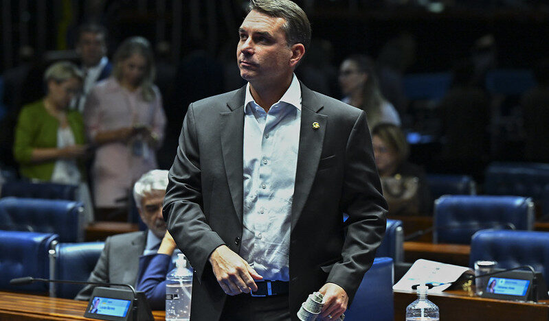 Flávio Bolsonaro em pé no plenário do Senado, segurando um microfone, com parlamentares e cadeiras ao fundo