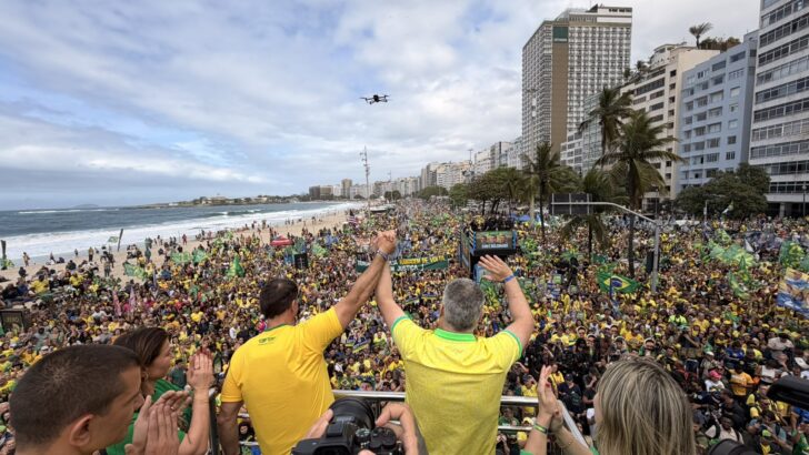Flávio Bolsonaro e Alfredo Gaspar aparecem de costas e com as mãos erguidas diante de uma multidão com bandeiras do Brasil na orla de Copacabana