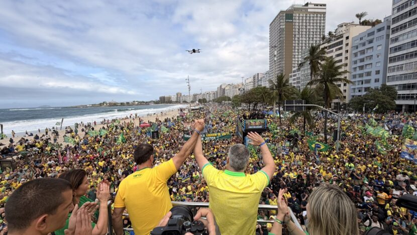 Flávio Bolsonaro e Alfredo Gaspar aparecem de costas e com as mãos erguidas diante de uma multidão com bandeiras do Brasil na orla de Copacabana
