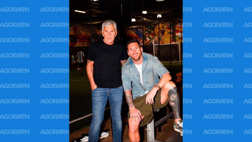 Jorge Messi em pé ao lado de Lionel Messi, que aparece sentado em um banco em ambiente esportivo