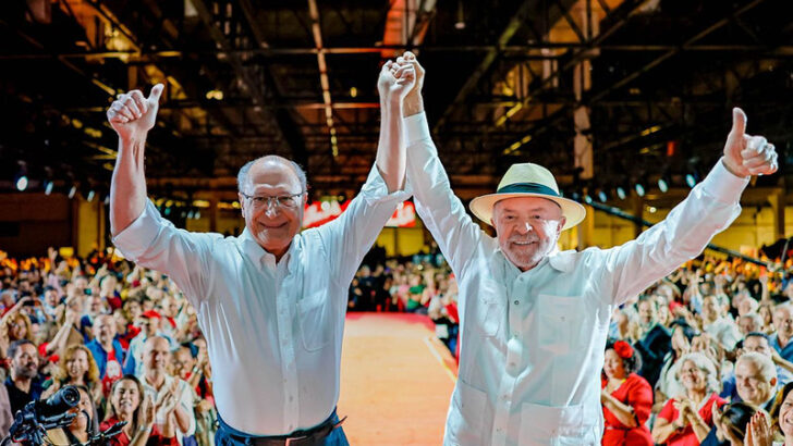 Lula e Geraldo Alckmin, vestidos de branco, levantam os braços diante de uma plateia durante convenção partidária
