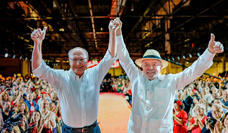 Lula e Geraldo Alckmin, vestidos de branco, levantam os braços diante de uma plateia durante convenção partidária