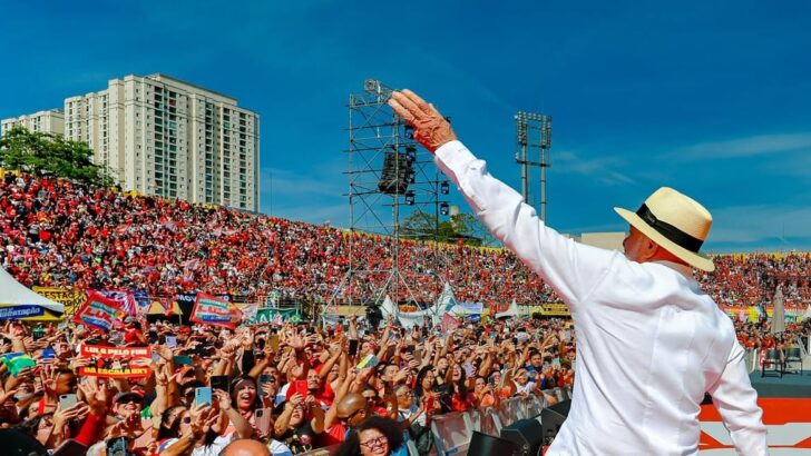 Lula, de camisa branca e chapéu, acena de costas para uma multidão vestida predominantemente de vermelho