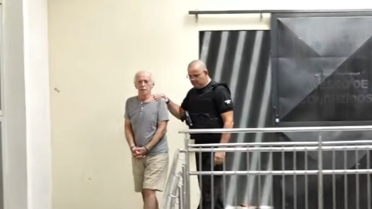 Marco Antônio Heredia Viveros, de camiseta cinza e bermuda, é conduzido por um agente de segurança em um corredor com grade metálica