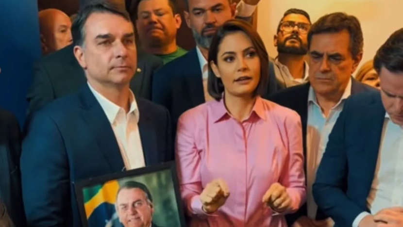 Michelle Bolsonaro, de camisa rosa, fala cercada por apoiadores enquanto Flávio Bolsonaro segura uma fotografia de Jair Bolsonaro