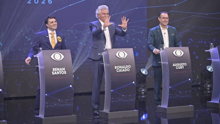 Renan Santos, Ronaldo Caiado e Augusto Cury em pé atrás dos púlpitos do debate da Band, com púlpitos vazios nas extremidades do palco
