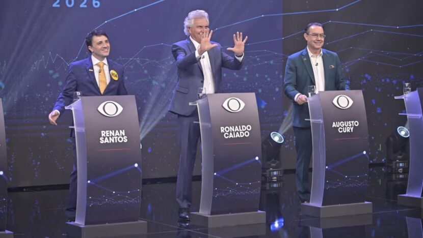 Renan Santos, Ronaldo Caiado e Augusto Cury em pé atrás dos púlpitos do debate da Band, com púlpitos vazios nas extremidades do palco
