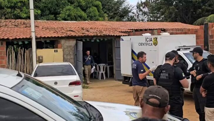 Agentes da Polícia Civil e da perícia estão reunidos diante de um imóvel, ao lado de viaturas e de um veículo da Polícia Científica