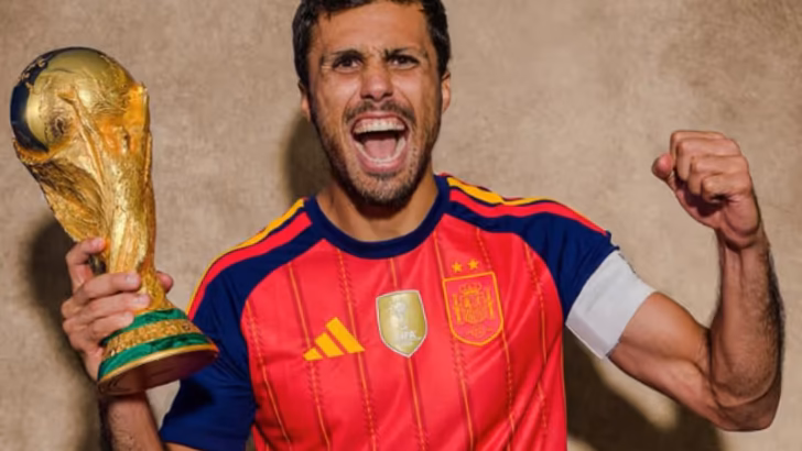 Jogador de futebol com uniforme da seleção da Espanha segura o troféu da Copa do Mundo e comemora com o braço erguido.