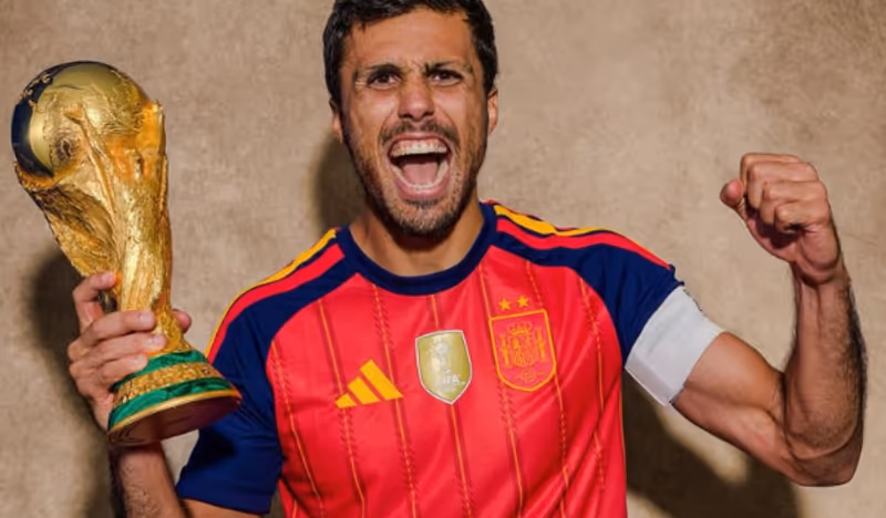 Jogador de futebol com uniforme da seleção da Espanha segura o troféu da Copa do Mundo e comemora com o braço erguido.
