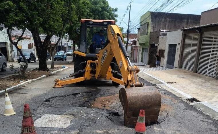 Retroescavadeira trabalha em trecho de rua com asfalto danificado e área sinalizada por cones