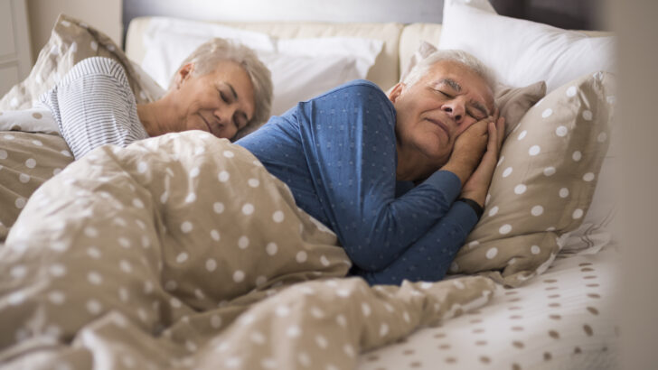 Duas pessoas idosas dormem deitadas em uma cama, cobertas e apoiadas em travesseiros