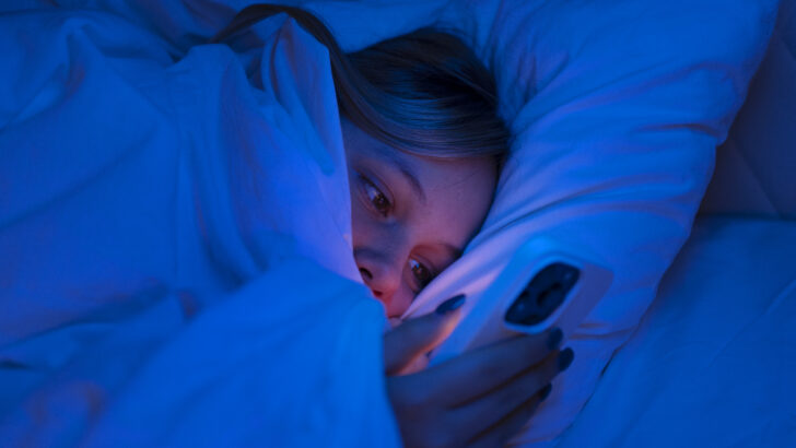 Mulher deitada na cama olha para a tela do celular durante a noite, com o rosto iluminado pela luz do aparelho