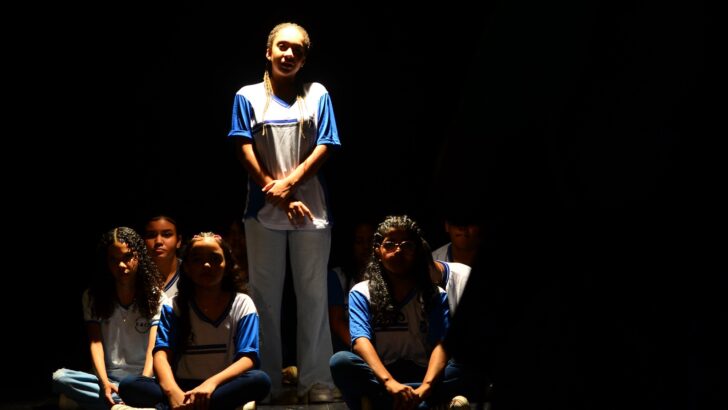 Jovens em cena teatral, com uma participante em pé e outras sentadas no palco