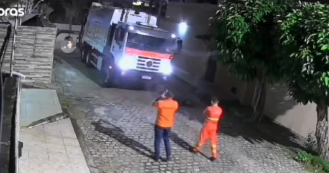 Caminhão de coleta de lixo parado em rua durante a noite, com dois trabalhadores próximos ao veículo