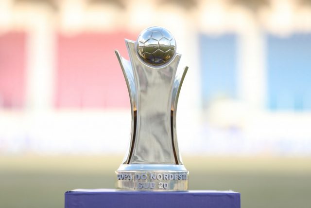 Troféu de competição de futebol sobre pedestal, com arquibancadas desfocadas ao fundo
