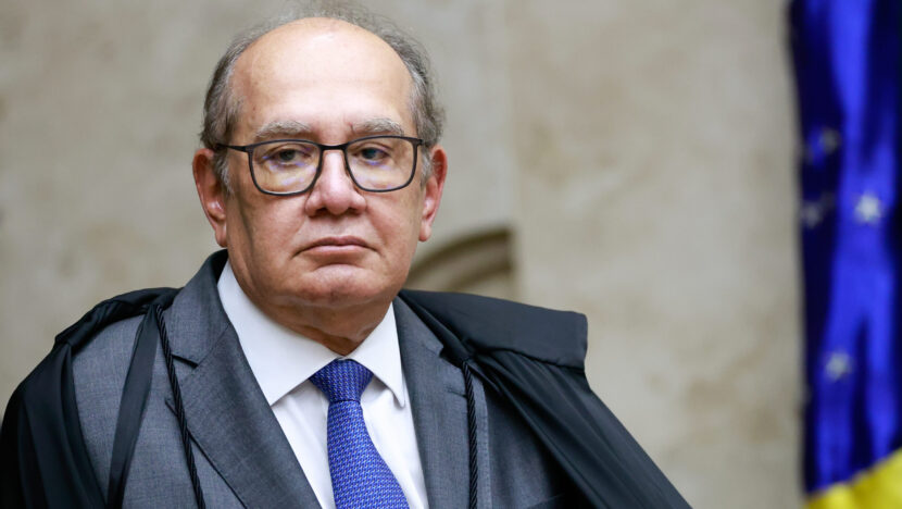 Ministro Gilmar Mendes, de toga e gravata azul, diante de uma bandeira do Brasil
