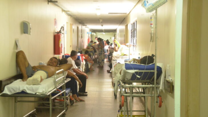 Pacientes em macas e cadeiras ao longo de corredor de unidade hospitalar