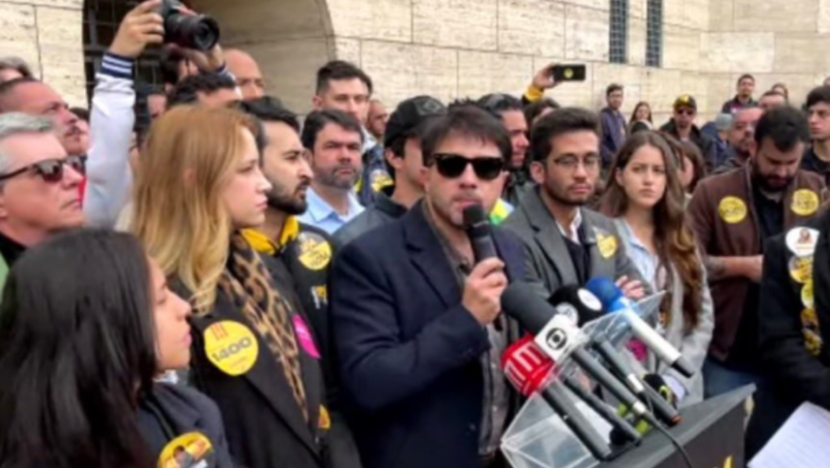Renan Santos, de óculos escuros e blazer, fala ao microfone diante de um púlpito com microfones de imprensa, cercado por apoiadores.