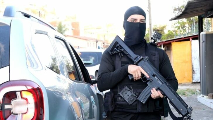 Policial com roupa preta e arma em frente a uma viatura durante ação policial