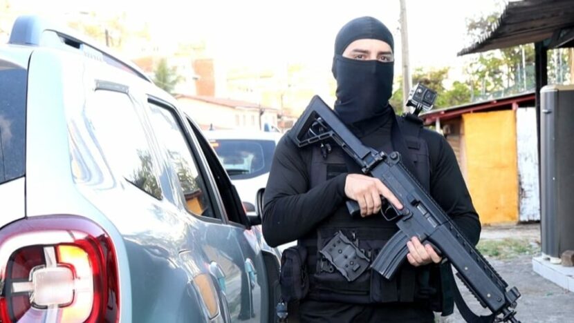 Policial com roupa preta e arma em frente a uma viatura durante ação policial