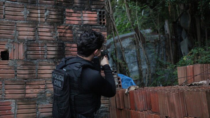 Policial civil com colete e arma durante ação em área cercada por paredes de tijolos