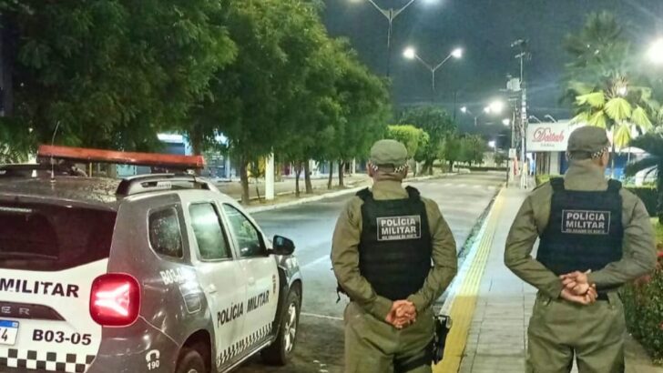 Dois policiais militares ao lado de uma viatura durante patrulhamento em uma via pública à noite