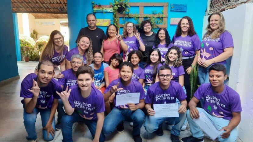 Grupo de estudantes uniformizados reunidos em uma biblioteca escolar, incluindo aluno em cadeira adaptada
