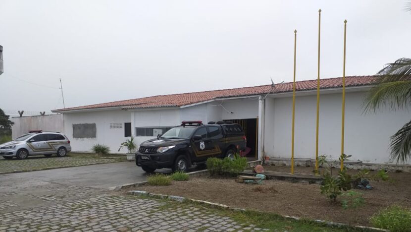 Área externa de unidade prisional com duas viaturas estacionadas em frente ao prédio