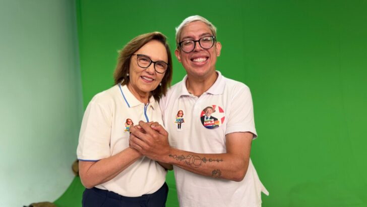 Duas pessoas posam juntas diante de um fundo verde, usando camisas brancas com adesivos de campanha