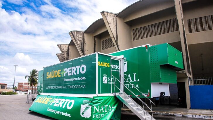 Carreta verde com a marca Saúde + Perto estacionada ao lado de um ginásio, com escada de acesso montada