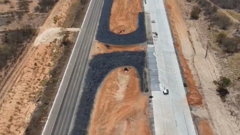 Vista aérea de pista nova de asfalto em construção ao lado da rodovia existente, em terreno de vegetação seca