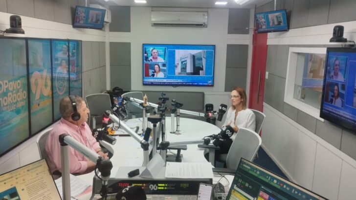 Estúdio de rádio com bancada, microfones e telas; uma mulher de blusa rosa usa fones de ouvido diante do microfone