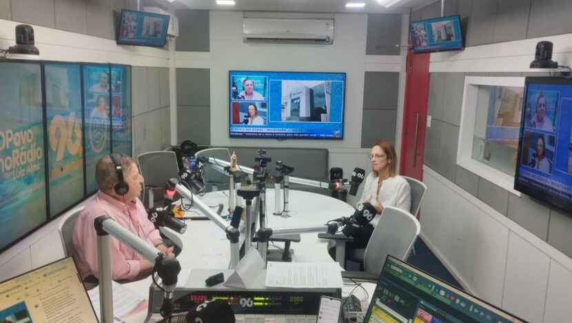 Estúdio de rádio com bancada, microfones e telas; uma mulher de blusa rosa usa fones de ouvido diante do microfone