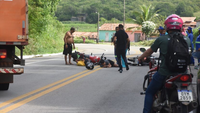 Motocicleta caída no meio da pista de uma rodovia, com pessoas em volta, um caminhão parado e outro motociclista passando