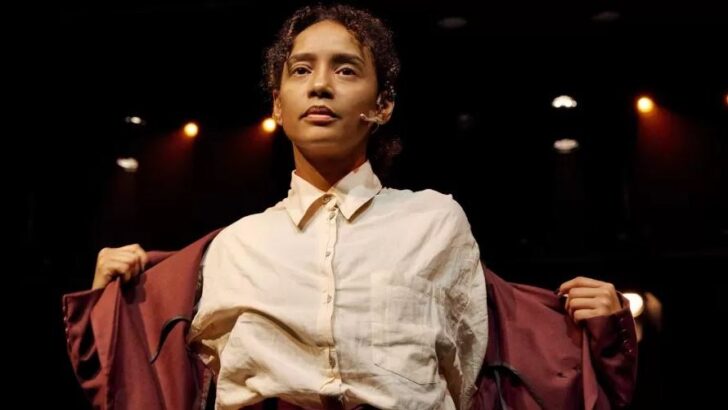 Atriz de cabelo curto e camisa clara abre um casaco vermelho-escuro em palco iluminado