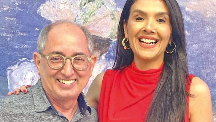 Homem de óculos e camisa clara e mulher de vestido vermelho sorriem diante de um painel com pintura em tons de azul