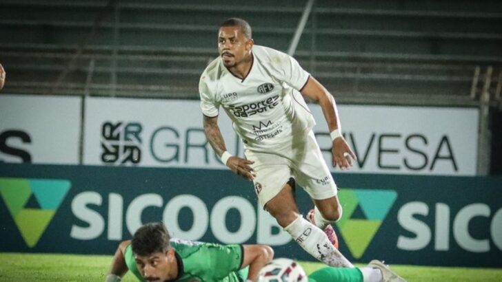 Atacante de uniforme branco avança com a bola enquanto um adversário de verde cai no gramado