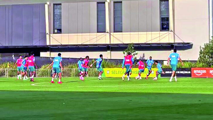 Jogadores em coletes de treino azuis e rosa disputam a bola em campo gramado, diante de banner da Seleção Brasileira