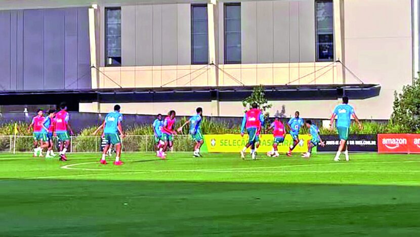 Jogadores em coletes de treino azuis e rosa disputam a bola em campo gramado, diante de banner da Seleção Brasileira
