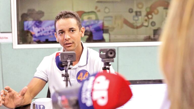 Homem de camisa polo branca gesticula diante de microfones de rádio e televisão em estúdio