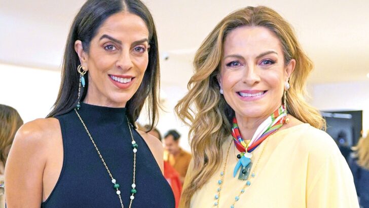 Duas mulheres posam lado a lado e sorriem em ambiente interno, uma de vestido azul-marinho e outra de conjunto claro