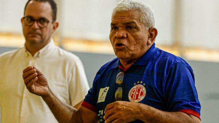 Dois homens conversam à beira da quadra; o da direita, de camisa do América, gesticula com as mãos