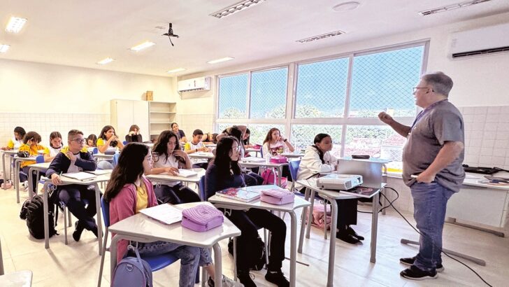 Estudantes sentados em carteiras de sala de aula acompanham a apresentação de um homem em pé ao lado da janela