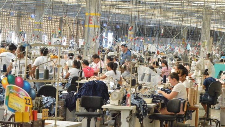 Galpão de confecção com dezenas de trabalhadores em máquinas de costura dispostas em fileiras