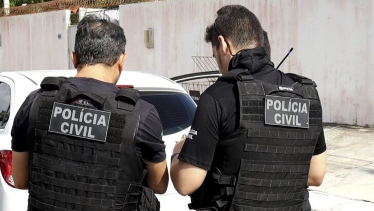 Dois agentes de colete da Polícia Civil, vistos de costas, ao lado de uma viatura