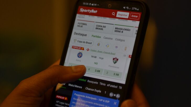 Mão segura um celular com a tela de um aplicativo de apostas esportivas aberta, em ambiente escuro