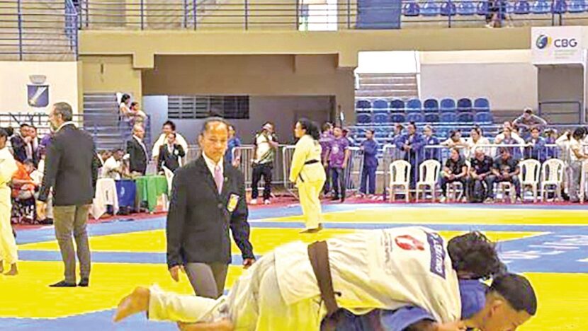 Atletas de judô disputam luta sobre o tatame de um ginásio, com árbitro e público ao redor